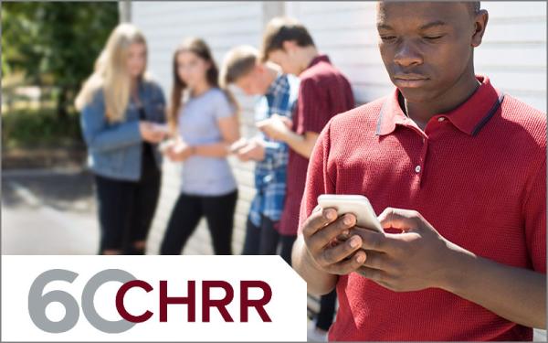 A black male teen looking at his phone, with peers behind him, blurred.
